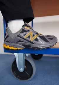 Noha opřená o modré kolečko kovového vozíku, na sobě černou a žlutou tenisku New Balance s bílou ponožkou a černými kalhotami.