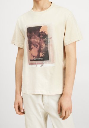 Mężczyzna w beżowej koszulce Calvin Klein z krótkim rękawem, ozdobionej abstrakcyjną brązowo-czarną grafiką i napisem z przodu, w połączeniu z jasnymi spodniami.