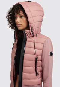 Gepolsterte rosa Jacke mit Kapuze, ausgestattet mit einem schwarzen Reißverschluss, seitlichen Taschen und strukturiertem Material. Mit einem Logo-Patch am Ärmel akzentuiert.