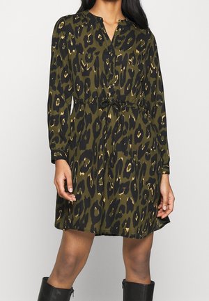 Kvinde iført en knap-op kjole med olivengrøn og sort leopardprint, med indsnøret talje, lange ærmer og sorte knæhøje støvler.