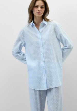 Joven mujer que lleva una camisa abotonada texturizada azul claro y pantalones a juego, de pie frente a un fondo blanco liso.