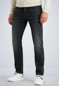 Muž oblečený v vyblednutých čiernych džínsoch a bielych teniskách stojí na svetlošedom podlahe so svetlým pozadím.