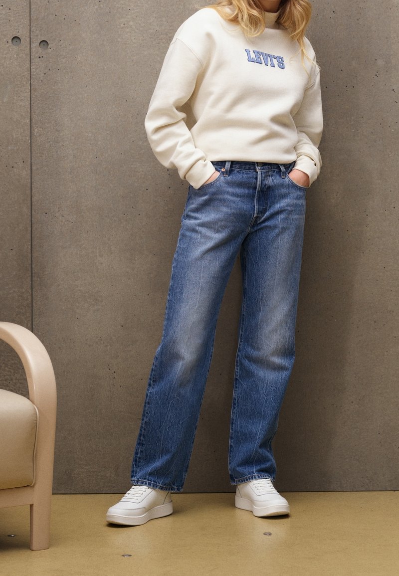Personne portant un sweat-shirt Levi's crème, un jean droit bleu et des baskets blanches, debout avec les mains dans les poches contre un mur en béton.