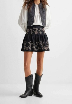 Czarna spódnica z haftem w kwiatowe wzory, w połączeniu z pikowaną ciemną kamizelką i białą długosleeve topem o fakturowanej powierzchni. Czarne kowbojki dopełniają stylizację.