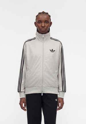 Person mit geflochtenem Haar trägt eine hellgraue Adidas Trainingsjacke mit schwarzen Streifen und Logo, kombiniert mit schwarzen Adidas-Hosen.