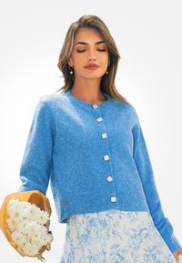 Blauwe gebreide cardigan met ronde halslijn en zes lichtgekleurde knopen. Model houdt een boeket witte bloemen vast, ingepakt in bruin papier.