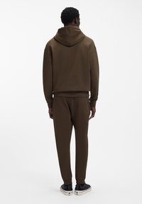 Barna kapucnis pulóver és hozzáillő nadrág puha anyagból; laza szabású, bordázott mandzsettákkal és kapucnival; fekete sportcipővel párosítva.