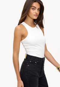 Witte geribbelde tanktop met brede bandjes, gecombineerd met hoge zwarte jeans met koperkleurige hardware en een leren label aan de achterkant.