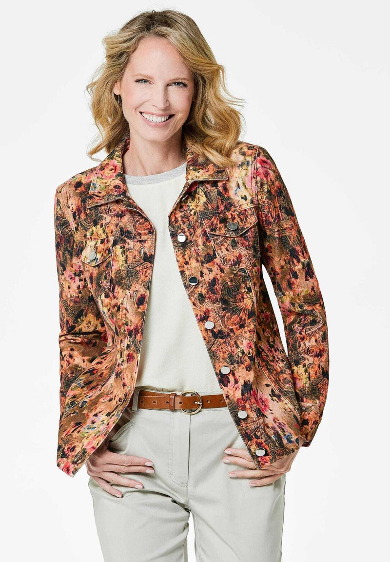 Kleurige spijkerjas met bloemenpatroon, voorzien van voorzakken, zilveren knopen en een aansluitend ontwerp, gedragen over een lichtgrijze top en beige broek.