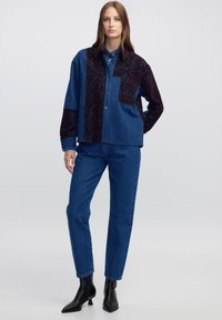 Spijkerjack met een donker, patroonstof aan één kant, voorzien van knopen, een kraag en een opgestikte zak. Gecombineerd met blauwe jeans en zwarte laarzen.