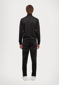 Black tracksuit with a stand-up collar, made from smooth fabric. Pants with side stripes and elastic cuffs, paired with white sneakers.