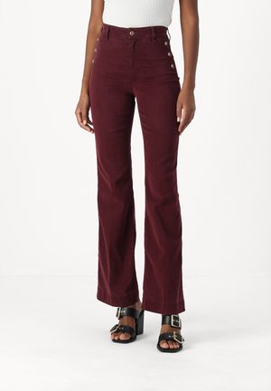Pantaloni a zampa d'elefante in corduroy color bordeaux con vita alta e dettagli in bottone dorato sui lati, abbinati a sandali neri con tacco quadrato.