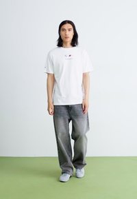 Hvid kortærmet T-shirt med logo, parret med løstsiddende grå denimbukser og grå sneakers. Ensfarvet baggrund med grøn gulv.