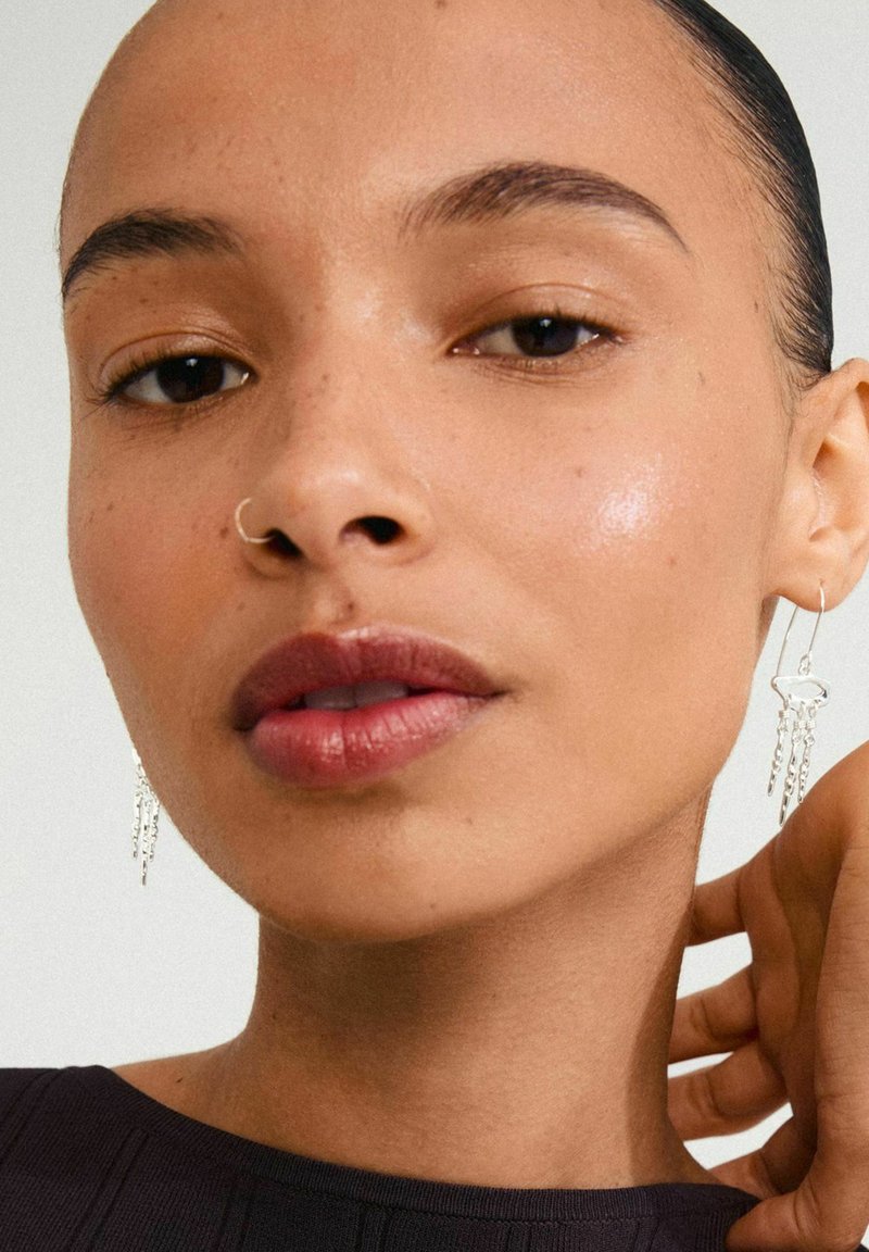 Pendientes de gota plateados con intrincados detalles en cadena, usados por una persona con piel suave y una blusa de tonos neutros, que presenta un maquillaje natural.