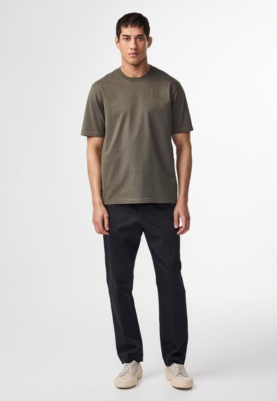 Oliven grøn bomulds T-shirt, kortærmet, rund halsudskæring, parret med mørke bukser og beige sneakers, stående foran en ensfarvet baggrund.