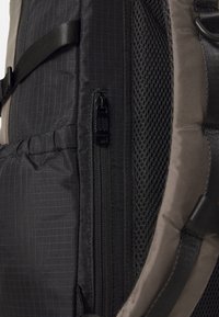 Crni platneni ruksak s teksturiranom površinom, opremljen džepom na patentni zatvarač, mrežastim panelom i podešavanim trakama. Vidljiv geometrijski uzorak.