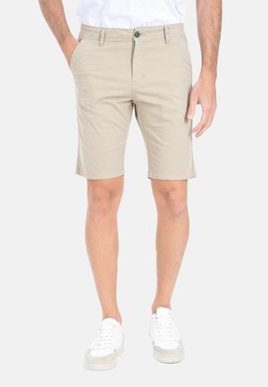 Uomo con pantaloncini beige fino al ginocchio, sneakers bianche e camicia bianca, in piedi con una mano in tasca contro uno sfondo semplice.