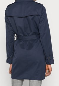 Manteau trench bleu marine avec taille ceinturée, épaulettes et poignets boutonnés. Texture lisse, longueur jusqu'aux genoux.