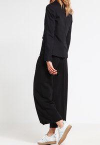 Blazer nero con una vestibilità aderente, abbinato a pantaloni neri a gamba larga. Footwear presenta accenti bianchi e verdi con una suola marrone.