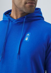 Niebieska bluza z kapturem i sznurkiem, wykonana z miękkiego materiału, z haftowanym logo Adidas i herbem Włoch na klatce piersiowej.