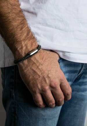 Čierny zapletený náramok so strieborným cylindrickým zapínaním na zápästí, umiestnený na bielom tričku a džínsovej pozadí.