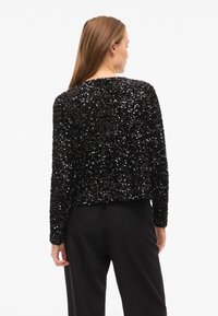 Črn pulover s svetlečimi sequini, dolgimi rokavi in okroglim izrezom, z teksturirano površino in skrajšano zasnovo, kombiniran s črnimi hlačami.