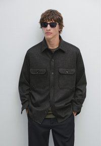 Camicia grigia scura con bottoni e due tasche frontali, un colletto e una consistenza morbida, indossata sopra una maglietta verde. Occhiali da sole neri completano il look.