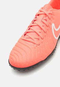 Fényes korall színű focicipő, texturált szintetikus felsőrésszel, fekete gumi talppal és fehér Nike logóval. Keskeny fűzős kialakítással a biztos illeszkedés érdekében.