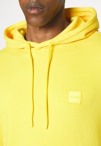 Gros plan d'une personne portant un sweat à capuche jaune vif avec cordons et un petit logo "BOSS" embossé sur la poitrine.