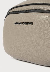 Pilkas tekstūruotas maišas su užtrauktuku, su juodu iškilniu „Armani Exchange“ logotipu priekyje. Dabartinė linija matoma palei užtrauktuką.