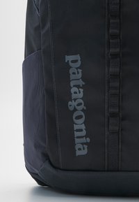Fekete szövet hátizsák texturált felülettel, nagy oldalsó zsebbel és szürke "Patagonia" logóval. Tartalmaz pántokat és minimális fém alkatrészeket.