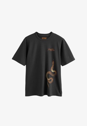 T-shirt noir à manches courtes et col rond avec un texte orange "Shinjuku Studios" et un graphisme détaillé d'un serpent enroulé sur le bas avant droit.