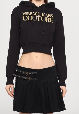 Femme portant un sweat à capuche court noir avec un texte doré "Versace Jeans Couture" et une jupe plissée noire avec deux sangles à boucle.