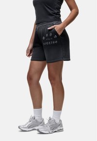 Shorts noirs avec un waistband élastique et des poches latérales, arborant le texte blanc "GOOD for NOTHING." Complétés par des chaussures de sport grises et des chaussettes blanches.