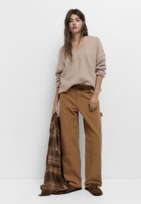 Beige strikket sweater, løs pasform; brune bomuldsbukser, lige ben; holder en mørk ternet jakke; brune sko med detaljeret syning.