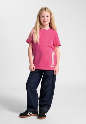 Ein junges Mädchen mit langen blonden Haaren steht da und trägt ein pinkes Hummel-T-Shirt, dunkle, weite Jeans und schwarze Sneakers mit weißen Chevron-Details.