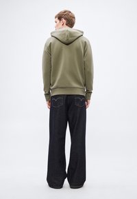 Felpa verde con cappuccio e cordino, polsini a coste, abbinata a jeans ampi in denim scuro con tasche posteriori e cuciture visibili.