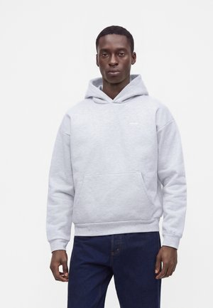 Giovane uomo con felpa grigio chiaro con tasca frontale e jeans blu scuro, in piedi davanti a uno sfondo bianco semplice.