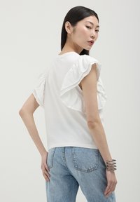 T-shirt bianca con maniche a volant, vestibilità morbida, abbinata a jeans azzurri. Il tessuto appare morbido, con una texture liscia e minimalismo nei dettagli.
