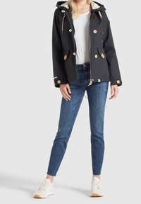 Veste imperméable noire avec des accents beiges, des boutons et une capuche à cordon, associée à un jean skinny bleu déchiré et des baskets blanches.