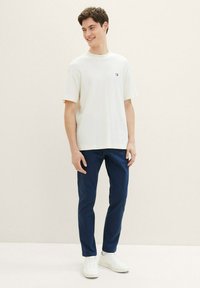 Vit kortärmad t-shirt med strukturerad tyg och liten logotyp, kombinerad med mörkblå byxor och vita sneakers. Enfärgad bakgrund.
