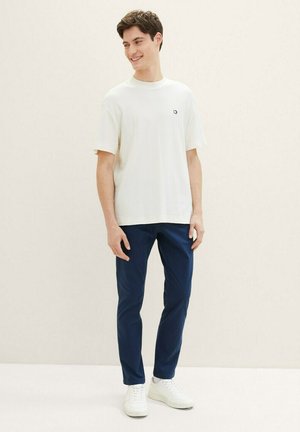 Weißes T-Shirt mit kurzen Ärmeln aus strukturiertem Stoff und kleinem Logo, kombiniert mit dunkelblauen Hosen und weißen Sneakers. Einfarbiger Hintergrund.