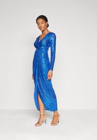 Μακρυμάνικο, σκούρο μπλε φορεματάκι με πούλιες, με σουρωμένο μπούστο και σκίσιμο στο μηρό. Διαθέτει ομαλή, γυαλιστερή υφή και εφαρμοστή σιλουέτα.