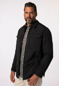 Schwarze Zip-Jacke mit zwei Brusttaschen, einem Kragen und einem leicht strukturierten Stoff, darüber trägt er ein graues Hemd mit Knöpfen.