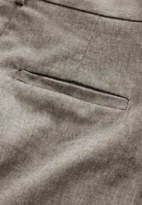 Gros plan d'une poche de pantalon en tissu gris avec une fente horizontale et des coutures visibles sur un matériau texturé.