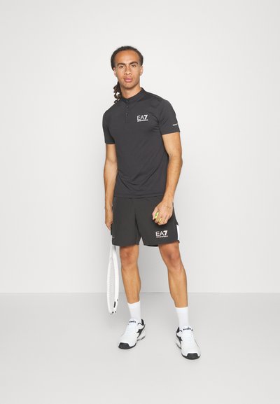 Polo noir avec col et manches courtes, associé à un short noir. Comprend le logo EA7. Le modèle tient une balle de tennis et une raquette.
