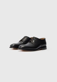 Pari di scarpe eleganti in pelle nera con lacci e tacco basso su uno sfondo bianco uniforme.