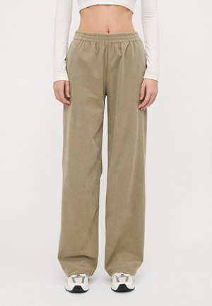 Beige Cordhosen mit weitem Bein und elastischem Bund, kombiniert mit weißen Sneakern und einem kurzen Langarmshirt, vor einem einfarbigen Hintergrund stehend.