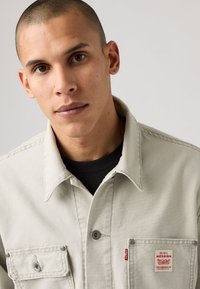 Chaqueta de mezclilla beige con cuello abotonado, bolsillo en el pecho y parche de logo rojo. Textura suave y detalles de costura sutil.