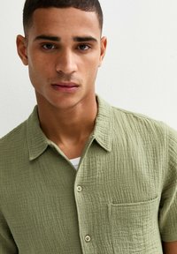 Camicia verde con texture, maniche corte, chiusura con bottoni e taschino sul petto. Realizzata in tessuto leggero con un motivo sottile.